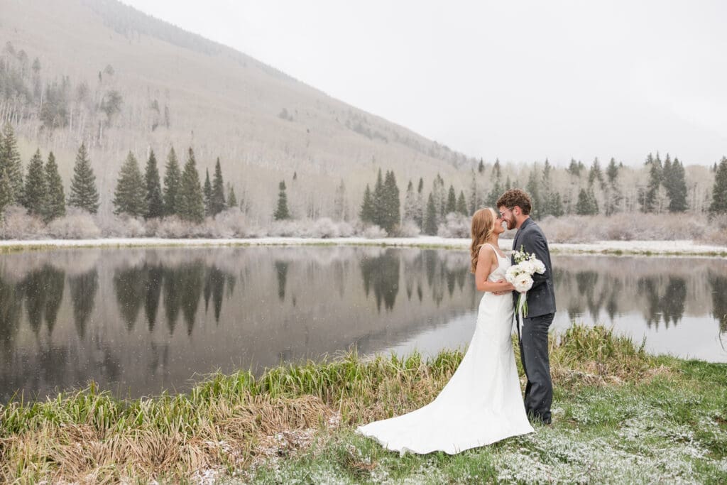 A bride and groom pose for a portrait against an alpine lake with the La Sal Mountains in the background